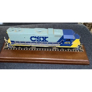 May include: A model train locomotive, featuring a light blue and navy blue color scheme with yellow accents. The CSX logo is prominently displayed on the side. The model sits on a wooden base with miniature tracks. The number 2678 is visible.