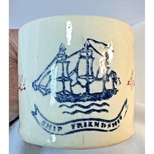 Op de afbeelding: Crèmekleurige keramische mok met een blauwe illustratie van een zeilschip. De woorden "SHIP FRIENDSHIP" staan op een banner onder het schip. Rode tekst staat aan de linker- en rechterkant van de mok.