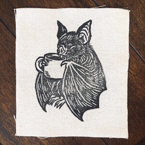 May include: A square off-white fabric patch featuring a black ink illustration of a bat holding a coffee mug with both wings. The bat is depicted with detailed fur texture and pointed ears, centered on the woven textile material with raw edges.