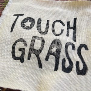 May include: A square piece of off-white canvas fabric featuring the black printed text TOUGH GRASS in a bold, hand-drawn style font. The letter O in TOUGH and the letter A in GRASS each contain a small, five-pointed star cutout design.