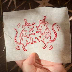 May include: A rectangular piece of light beige fabric featuring a red embroidered design of two mice standing on their hind legs. Each mouse wears a small party hat, and the illustration includes curved lines around them to suggest movement.