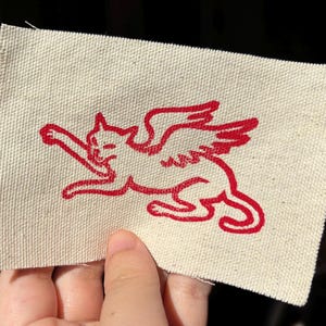 May include: A rectangular off-white fabric patch featuring a red embroidered graphic of a winged cat in a leaping pose. The design shows the cat with extended wings and a long tail, rendered in a bold red thread against the textured canvas material.