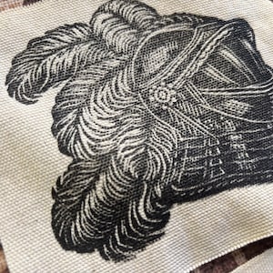 May include: A square piece of off-white fabric featuring a detailed black ink illustration of a historical helmet adorned with large, flowing feathers. The print displays intricate line work and shading, capturing the texture of the plumes.