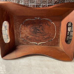 Può includere: Vassoio di legno con bordi curvi e manici ritagliati. Il vassoio presenta un centro decorativo con le parole "Give Us This Day Our Daily Bread" in un carattere decorativo. Il vassoio è di colore marrone caldo.