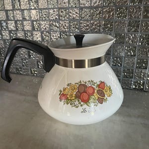May include: White ceramic coffee pot with a black handle and lid. The pot features a decorative floral and fruit design with the text "Le Roi". A silver band encircles the middle of the pot.