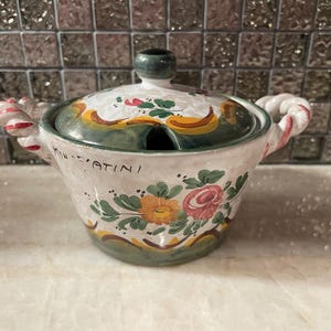 Op de afbeelding: Een kleine, handbeschilderde keramische pot met een deksel. De pot is wit met bloemmotieven in roze, geel en groen. Hij heeft twee gedraaide handvatten en een groene knop op het deksel. De woorden "MON-FATINI" staan op de zijkant.