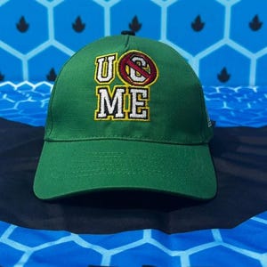 Op de afbeelding: Groene baseballcap met de tekst "U Can't See Me" geborduurd in geel, rood en wit. De cap heeft een gebogen klep en een zwarte knoop. Gemaakt van duurzaam materiaal.