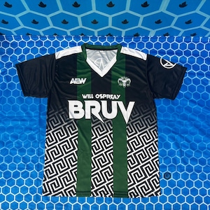 May include: A black and green V-neck jersey featuring the text AEW, WILL OSPREAY, and BRUV on the front. The shirt has white shoulder panels, a white geometric Greek key pattern along the bottom, and a white circular logo on the left sleeve.
