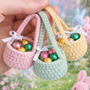 May include: Three miniature crochet Easter baskets in pastel pink, green, and yellow, each filled with foil-wrapped chocolate eggs. The baskets have small handles and white ribbon bows. The background is a blurred, colorful spring scene.