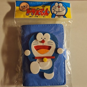 May include: Blue wallet featuring the cartoon character Doraemon. The wallet has a large Doraemon figure on the front with white, red, and yellow accents. The wallet is packaged in a clear plastic bag with a yellow and blue header.