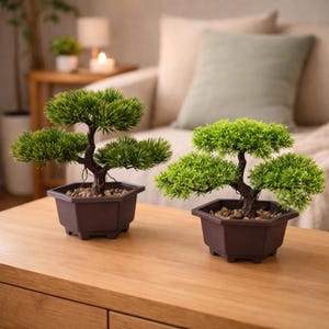 May include: Two artificial bonsai trees in dark brown hexagonal pots. The trees have green foliage and brown trunks, with small pebbles at the base. They are placed on a light brown wooden surface, with a sofa in the background.