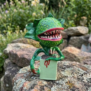 Könnte beinhalten: Eine handbemalte grüne Keramikskulptur einer fleischfressenden Pflanze mit einem großen, offenen Maul, das scharfe Zähne zeigt. Die Pflanze befindet sich in einem hellgrünen Topf mit einem roten Symbol. Die Skulptur steht auf einer Steinoberfläche.
