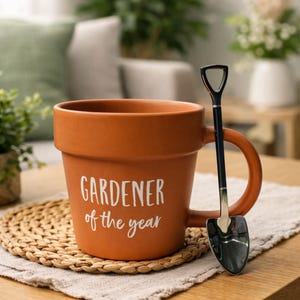 Könnte beinhalten: Eine terrakottafarbene Tasse in Form eines Blumentopfs mit dem weißen Schriftzug "GARDENER of the year". Neben der Tasse liegt ein kleiner, schaufelförmiger Löffel. Die Tasse steht auf einem gewebten Untersetzer.