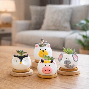 May include: Four ceramic animal-shaped planters with succulents. The planters are white with painted details: an owl, a bear, a cow, and an elephant. Each planter sits on a small, round wooden base. The background includes a wooden table and a blurred living room.