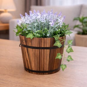 以下が含まれることがあります： ラベンダーの花と緑で満たされた木製の樽型のプランター。プランターは、黒い金属バンドが付いた濃い茶色の仕上げで、木製のテーブルの上に置かれています。ラベンダーの花は薄紫色です。