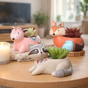 May include: Three animal-shaped planters, a fox, a raccoon, and a fox, each holding a succulent plant. The fox is orange, the raccoon is white and grey, and the fox is pink. Each planter has a Native American design.
