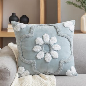 May include: A square, pale blue cushion featuring a large white flower design. The flower and border have a textured, raised appearance. The cushion is on a grey sofa with a white blanket.