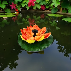 Puede incluir: Una fuente de flor de loto naranja y amarilla vibrante flota en un estanque. La fuente tiene una boquilla negra en el centro y descansa sobre una gran hoja de nenúfar verde. El estanque está rodeado de follaje verde y flores rosas.