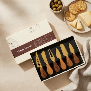 May include: A cheese knife set in a gift box. The set includes six knives with gold blades and wooden handles. The box is white with a brown stripe and the words "Cheese knife set". Olives, crackers, and cheese are also visible.