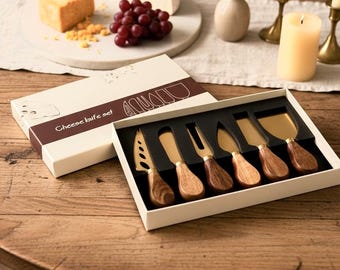 May include: A cheese knife set in a white box with a brown band labeled Cheese knife set. The set includes six gold-toned metal knives with wooden handles, featuring various blade shapes for cutting and serving different types of cheese.