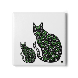 Op de afbeelding: Vierkante, witte keramische tegel met twee zwarte katten silhouetten. De katten zijn versierd met witte bloemen en groene bladeren. De tegel heeft een glanzende afwerking en een klein logo rechtsonder.