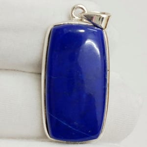 May include: A rectangular pendant with a vibrant blue stone set in a silver-colored frame. The stone has a smooth, polished surface and a small loop at the top for a chain.