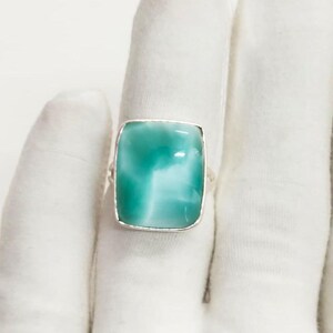 May include: A square-cut, turquoise-colored gemstone ring with a silver band. The gemstone has a marbled appearance with white and light blue swirls. The ring is set on a white background.