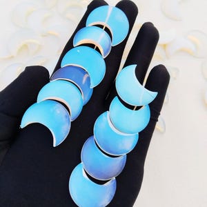 May include: Two pairs of earrings, each with a sequence of crescent moon shapes in shades of blue. The earrings are set against a black backdrop, with white floral accents in the background.