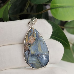 May include: A teardrop-shaped pendant with a polished blue and brown stone set in a silver-colored metal frame. The stone has a marbled appearance with streaks of blue, brown, and yellow. The pendant is displayed against a white background.