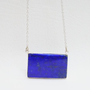 May include: A rectangular lapis lazuli pendant necklace. The deep blue stone is set in a silver-toned metal frame and hangs from a delicate silver chain. The necklace is displayed against a white background, highlighting the vibrant color of the gemstone.