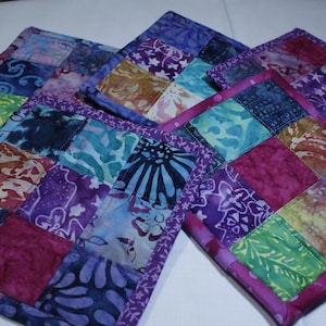 May include: A collection of handmade square fabric coasters. Each coaster is a patchwork of colorful batik fabric squares in shades of purple, blue, green, and orange. The edges are finished with a coordinating purple binding.