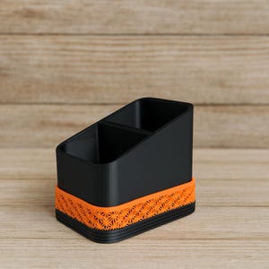 Puede incluir: Un organizador de escritorio negro con dos compartimentos. El organizador tiene una banda decorativa naranja con un patrón de estrella alrededor de la base. El organizador está hecho de plástico y se encuentra sobre una superficie de madera.