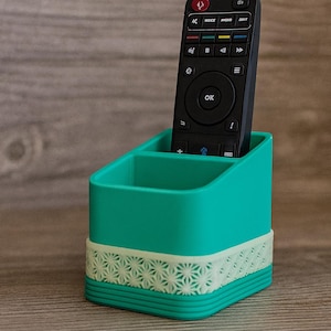 Può includere: Un porta telecomando turchese con una fascia a motivi bianchi. Un telecomando nero è posto all'interno. Il supporto è in plastica e poggia su una superficie di legno. Il telecomando ha un pulsante "OK".