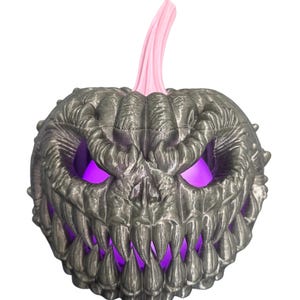 May include: A metallic gray Halloween pumpkin with a menacing face and a pink stem. The pumpkin's eyes and mouth glow with a bright purple light, creating a spooky effect. The design features sharp teeth and a textured surface.