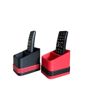 May include: Two remote control organizers, one black and one red, each with a geometric patterned base. The organizers have two compartments and hold a black remote control. The design is modern and functional.