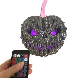 May include: A spooky Halloween pumpkin decoration with a menacing face, glowing purple eyes and mouth, and a pink stem. A remote control is held nearby to change the light colors. The pumpkin is dark gray.