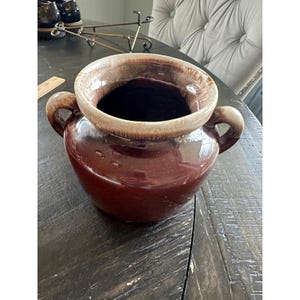 Puede incluir: Un jarrón de cerámica marrón con dos asas. El jarrón tiene una abertura ancha con un borde marrón claro. El exterior es de color marrón oscuro brillante. El jarrón está sobre una mesa de madera oscura.