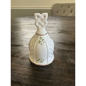 Puede incluir: Campana de cerámica blanca con borde dorado y asa de nudo celta. La campana presenta diseños de tréboles verdes y patrones de hojas en relieve. Un artículo decorativo para la decoración del hogar.
