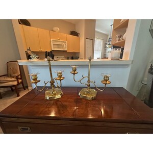 Puede incluir: Dos candelabros de latón a juego con un acabado en tono dorado pulido. Cada candelabro cuenta con un poste central vertical con dos brazos curvos ajustables que se extienden hacia afuera, rematados con arandelas de vidrio transparente.