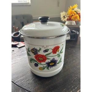 May include: A white enamel pot with a matching lid, featuring a colourful floral pattern of red, purple, and yellow flowers with green leaves. The pot has two black side handles and a black knob on the lid, with a silver-toned metal rim.