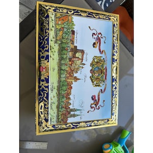 May include: Rectangular decorative tray with a detailed illustration of a European town, featuring buildings, trees, and a river. The tray has a gold-coloured frame with blue and gold decorative accents. The illustration includes text in a foreign language.