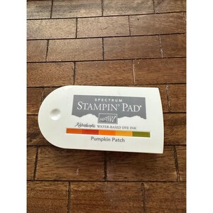 Peut inclure: Un tampon encreur blanc Stampin' Pad avec le texte "SPECTRUM STAMPIN' PAD" en gris. En dessous, un dégradé de couleurs orange, jaune et vert. Le tampon encreur est étiqueté "Pumpkin Patch" en orange foncé.