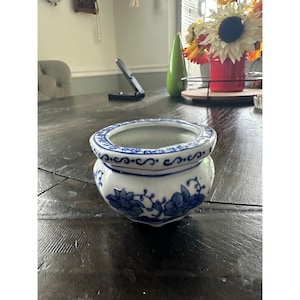 Könnte beinhalten: Ein kleiner, runder Übertopf aus weißer Keramik mit einem blauen Blumenmuster auf der Außenseite und einem dekorativen blauen Schnörkeldesign am dicken, abgerundeten Rand. Das schalenförmige Gefäß hat eine glatte, glänzende Oberfläche.