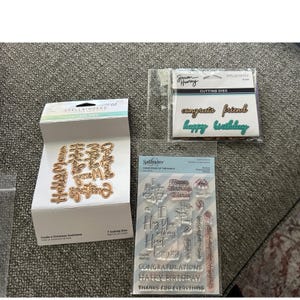 Puede incluir: Una colección de suministros para manualidades, que incluye troqueles y sellos transparentes. Los troqueles son dorados y presentan sentimientos navideños. Los sellos incluyen mensajes de cumpleaños y felicitaciones. Los artículos están empaquetados en plástico transparente.