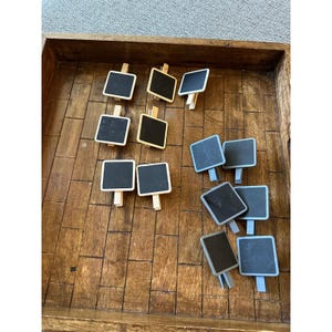 May include: A collection of small square chalkboard signs with clips. Five signs feature natural light-coloured wooden clips, while five signs feature light blue painted wooden clips. Each sign has a black surface for writing with chalk.