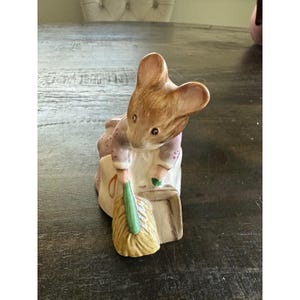 May include: A ceramic figurine of a brown mouse wearing a purple jacket and white apron. The mouse is depicted in a forward-leaning posture, holding a green-handled broom and sweeping a small pile of yellow dust into a tan dustpan.