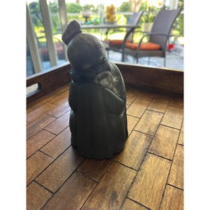 Può includere: Una scultura grigio scuro di una figura seduta, forse una persona anziana, con una postura curva. La figura indossa un abito lungo e ha uno chignon in cima alla testa. La scultura è su un vassoio di legno.