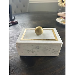 May include: A rectangular white marble box with grey veining, featuring a gold-colored rectangular border on the lid and a centered, spherical gold-colored knob handle. The box has a smooth, polished finish and a clean, geometric design.