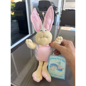 Könnte beinhalten: Eine Bunnybees-Plüschpuppe mit einem rosa-blau gestreiften Outfit, langen rosa Ohren und einem Etikett mit der Aufschrift "A Flying, Furry Tale". Die Puppe hat ein cremefarbenes Gesicht und Gliedmaßen und hält die Arme hoch.