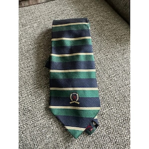 Puede incluir: Una corbata con rayas horizontales en azul marino, verde bosque y finas líneas color crema. La corbata incluye un pequeño logotipo bordado en forma de escudo cerca de la punta inferior, que muestra un león rojo y una cinta debajo.